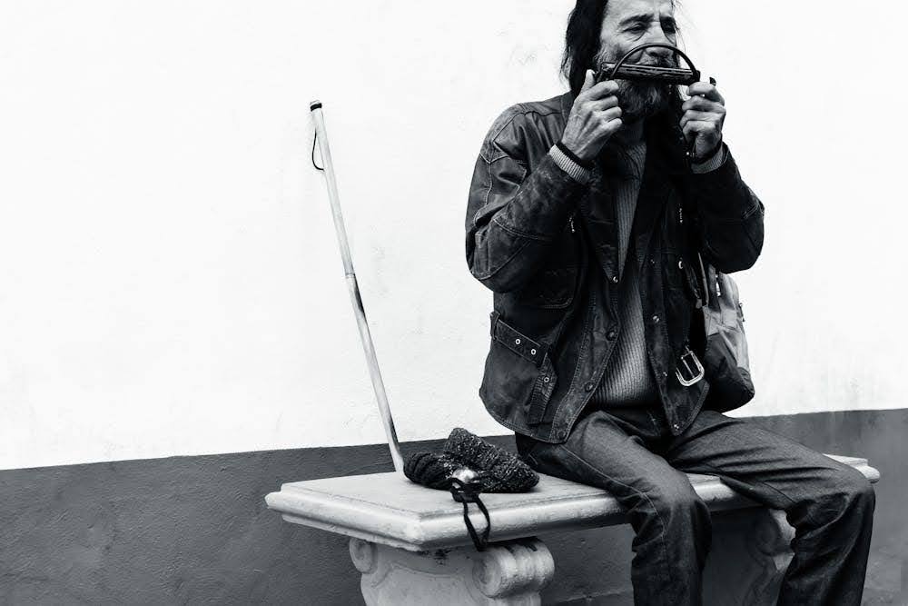 A black and white photo of a street musician playing a harmonica while sitting on a bench